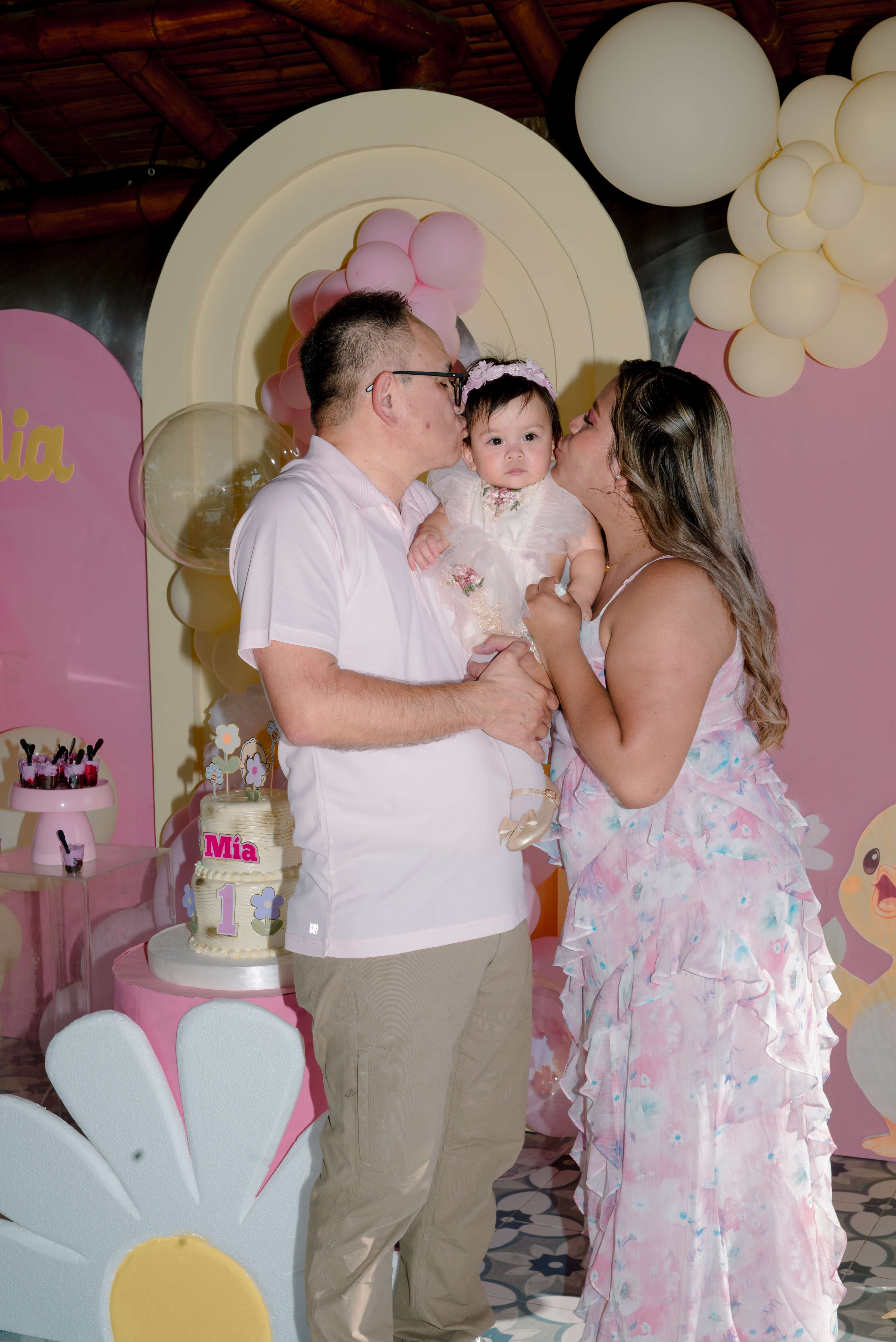 Papá y mamá besando a Mía en su primer cumpleaños frente a la mesa dulce decorada en rosa y crema con el pastel de un año en Las Marías Playas